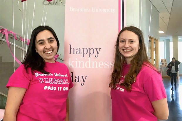 Two students in bold pink Kindness day t-shirts pose with the Brandeis Kindness Day banner in the Shapiro Campus Center.